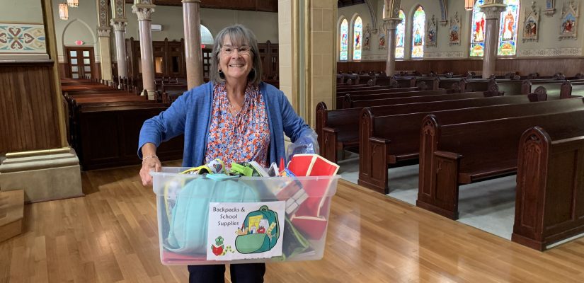 A smiling woman stands holding a container labeled Backpack & School Supplies