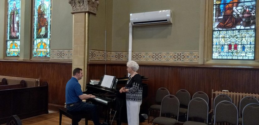A man playing the piano in church while a woman stands beside the piano.