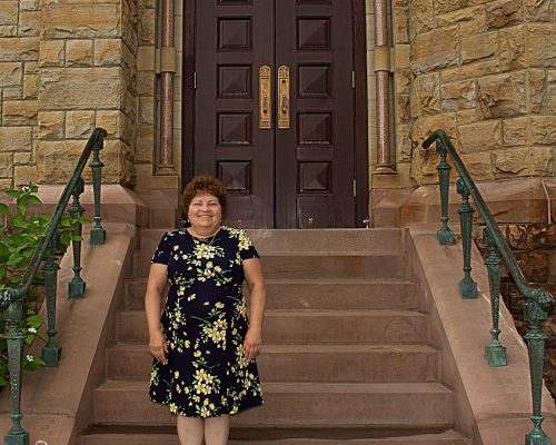 A smiling woman stands at the bottom of stone steps leading up to a church