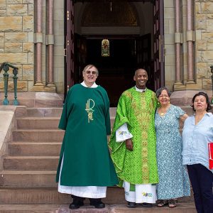 A Priest and Deacon stand with 2 women on the church steps.