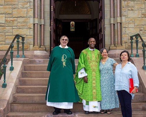 A Priest and Deacon stand with 2 women on the church steps.