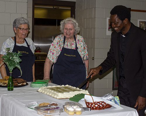 Priest cutting cake while 2 smiling ladies look on