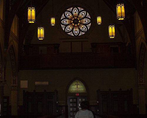 A woman is walking down the aisle toward the door with stained glass windows and lights illuminating the path
