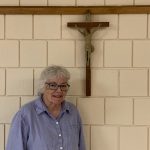 A smiling woman is standing in front of a crucifix