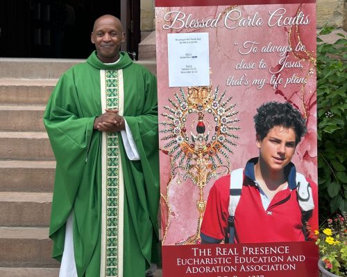A Priest stands next to a poster: Vatican International Exhibit. The Eucharistic Miracles of the World. In Parish Hall