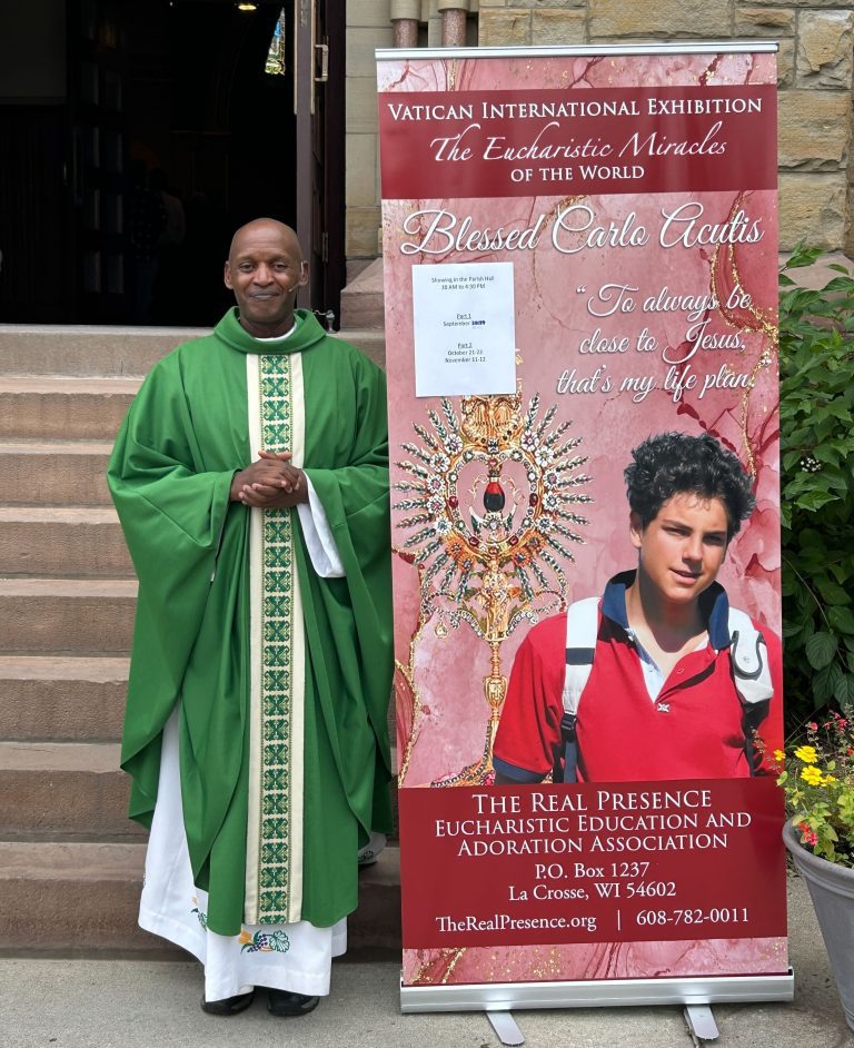 A Priest stands next to a poster: Vatican International Exhibit. The Eucharistic Miracles of the World. In Parish Hall