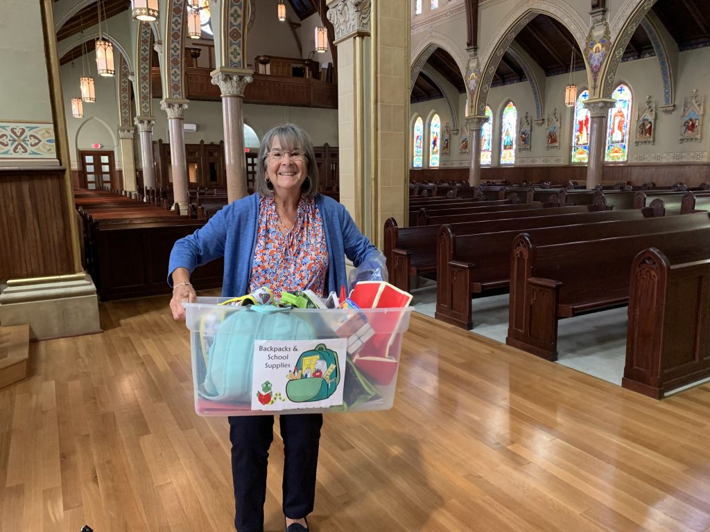 A smiling woman stands holding a container labeled Backpack & School Supplies