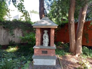 Virgin Mary statue surrounded by brick and stone with Ave Maria Gratia Plena marker among a garden.